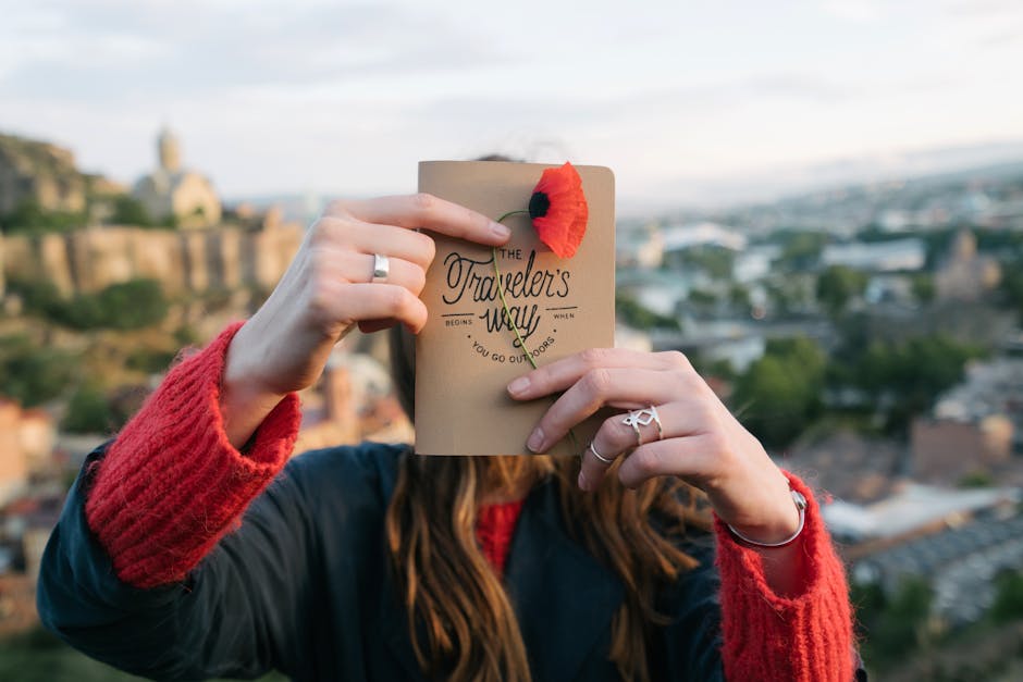 Woman holding a travel-themed booklet with a poppy in an outdoor scenic setting.